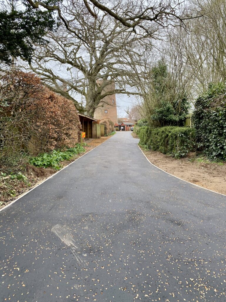 A newly laid asphalt private road with a smooth asphalt surface, surrounded by trees and bushes on either side, leading to buildings in the distance including aylsham windmill on a cloudy day.
