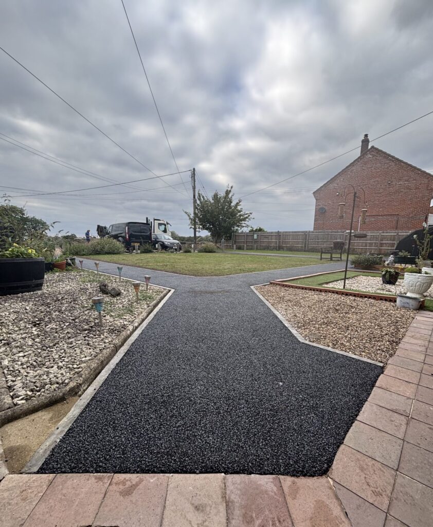 A newly laid black permeable asphalt footpath with concrete kerb edging on a cloudy day with a house and garden in the background.
