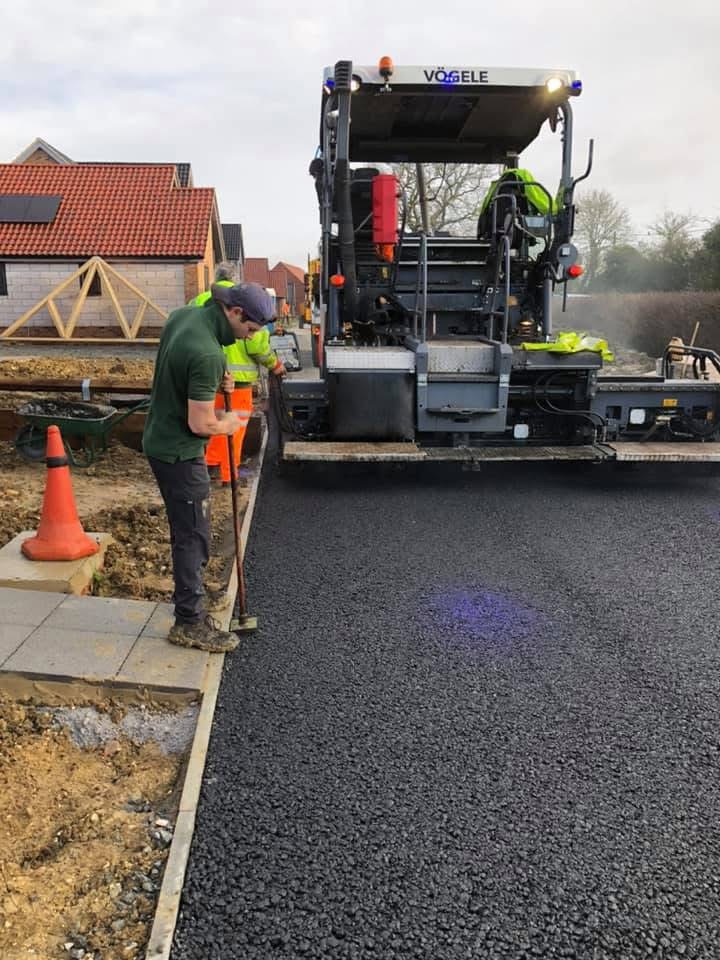 Workers lay tarmac on housing estate with paving machine nearby on cloudy day