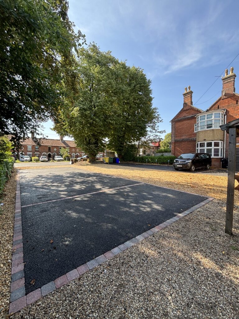 A newly laid tarmac car park with brick edging, surrounded by gravel and trees, with a house and cars in a nursery car park on a sunny bright day