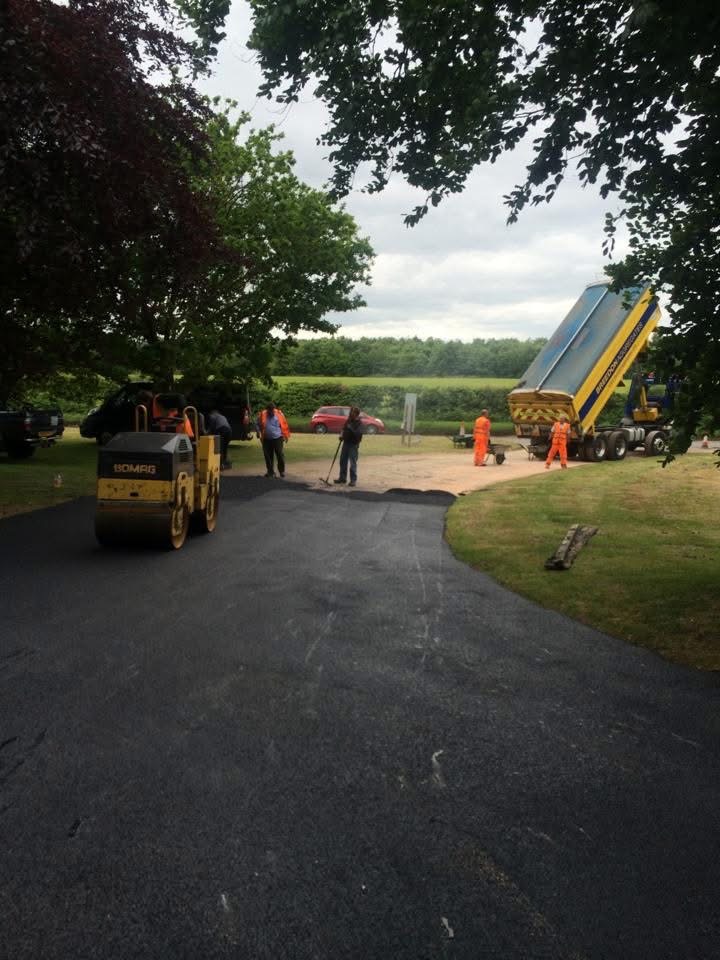 Workers in orange and blue clothing lay asphalt on private roadway with yellow roller and truck nearby outdoors
