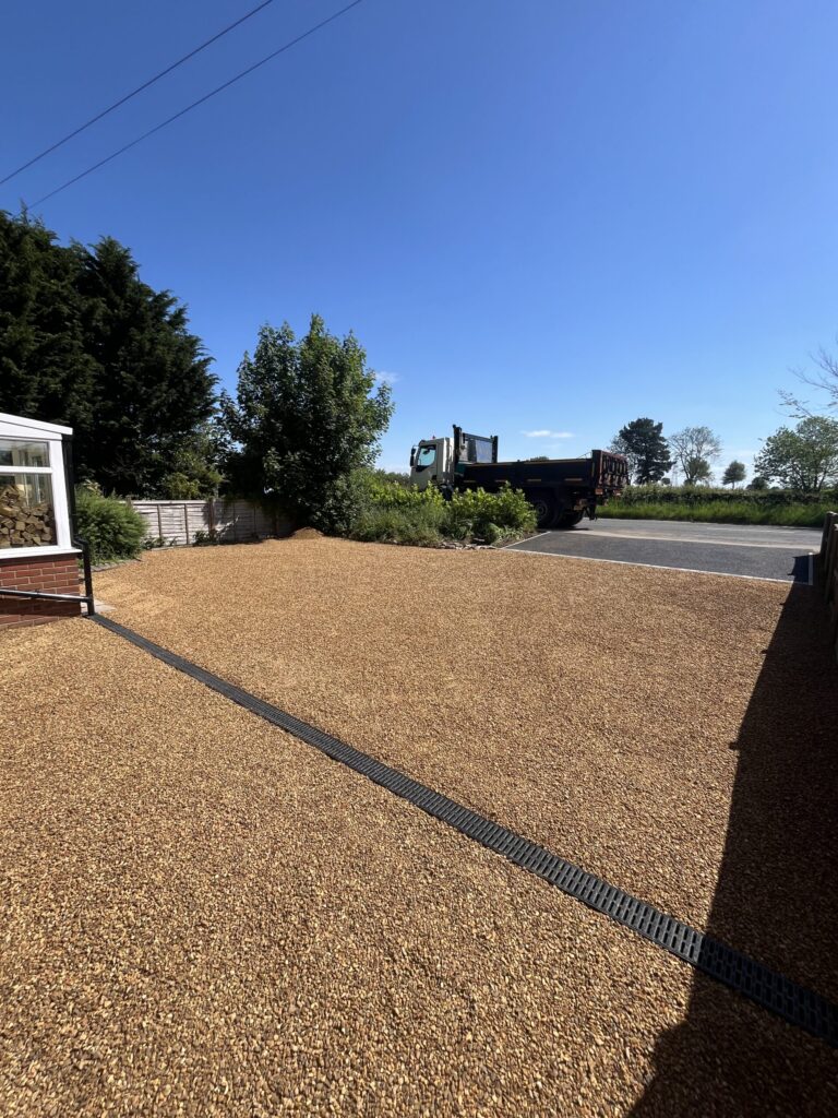 A large area of tar and chip surfacing on a driveway in norfolk with a tarmac truck on road, brown gravel surface, green trees, blue sky